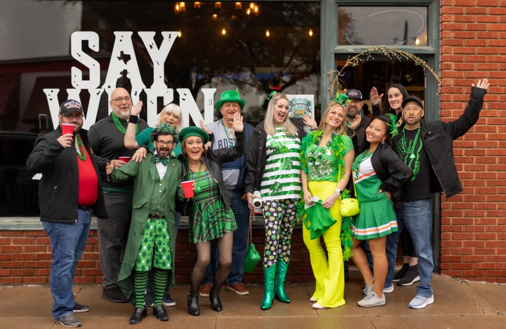 A group of twelve adults dressed in green celebrate out front of a brewery.
