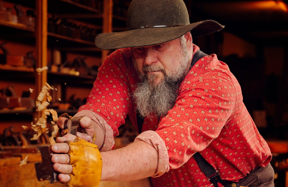 Bearded man is shaving wood intently to create hand-made furniture.