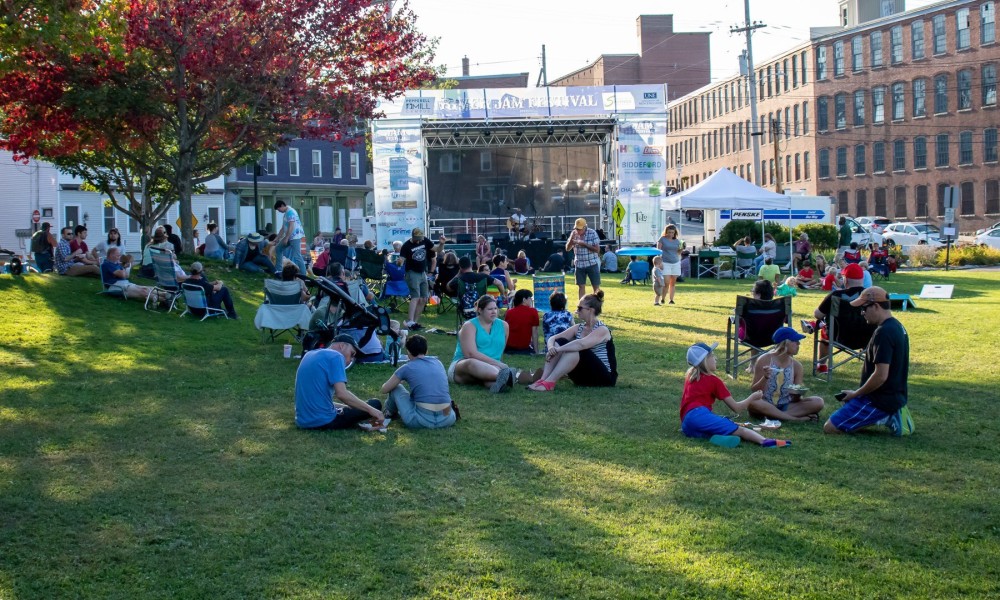 People gather in small groups on a shady grass lawn in front of a concert stage.