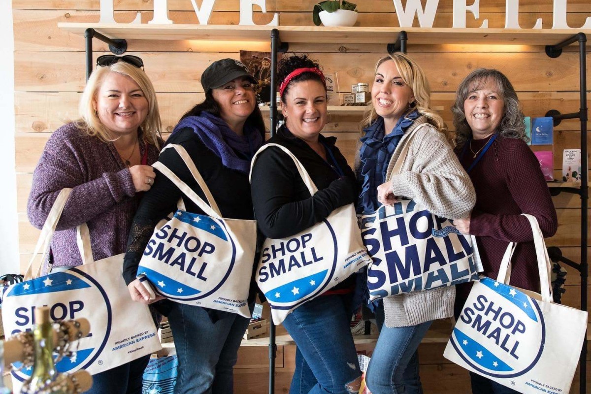 A group of women pose with a group of "Shop Small" tote bags.