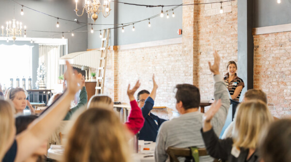 People sitting around tables raise their hands; in the background a speaker holds a microphone.