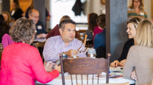 People sit around a table; in the center, a man gestures while speaking.