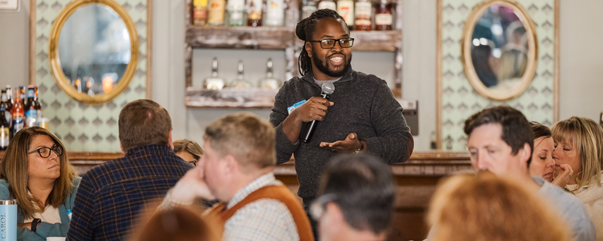 A man stands while speaking into a microphone; he is surrounded by people sitting and listening intently.