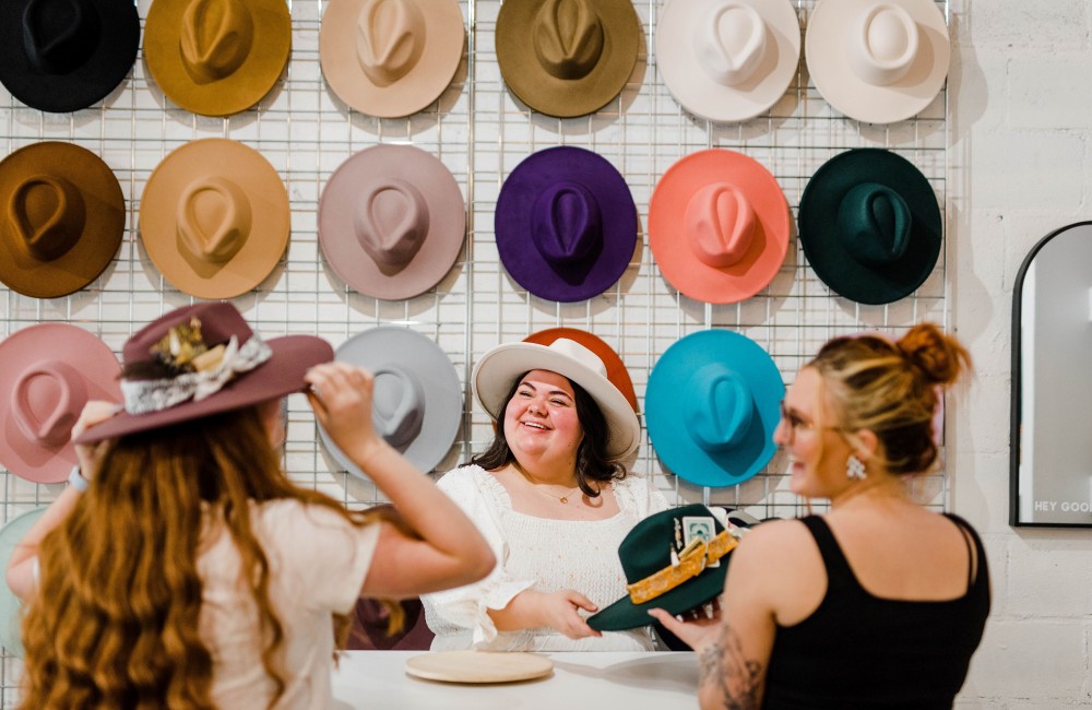 Three women are trying on hats in front of a large wall of hats.