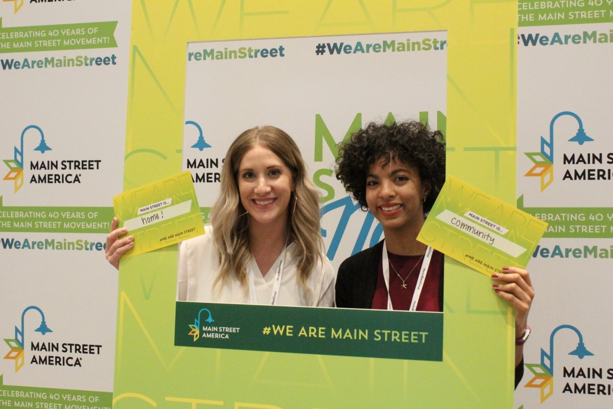 Two attendees at the Main Street Now Conference stand in front of the We Are Main Street campaign step and repeat.