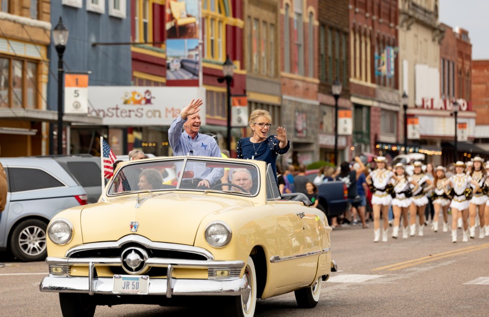 A woman and a man wave from the open top of a classic convertible during a parade.
