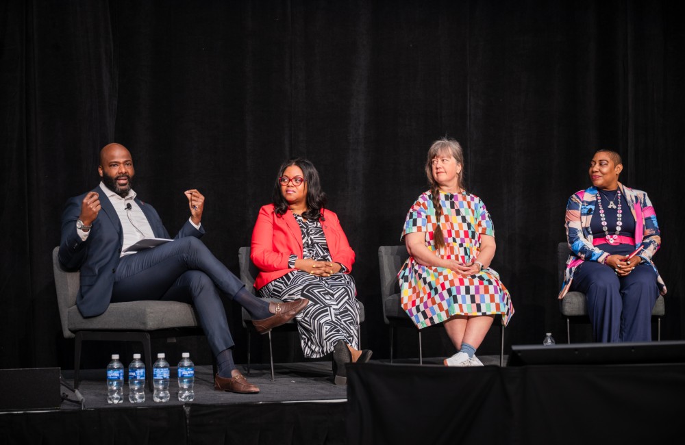 Four people sit in chairs on a stage; the three people on the right look at the person on the left who is speaking.