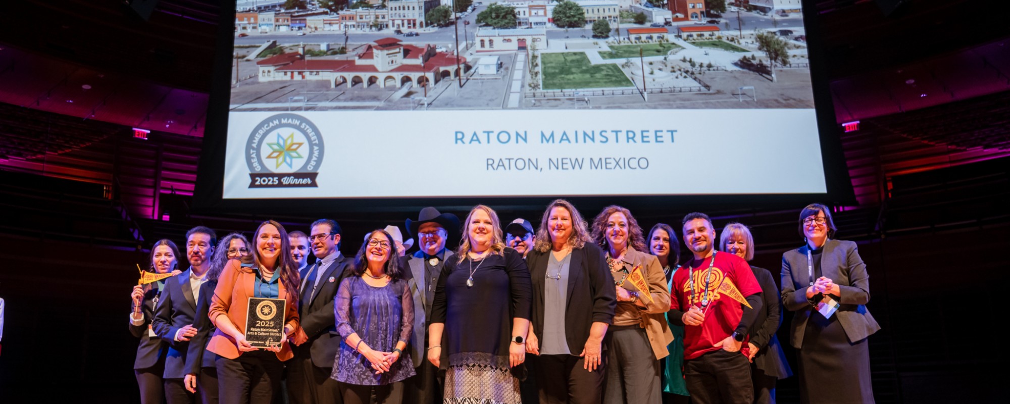 Large group on stage in front of a screen that reads "RATON MAINSTREET - RATON, NEW MEXICO."