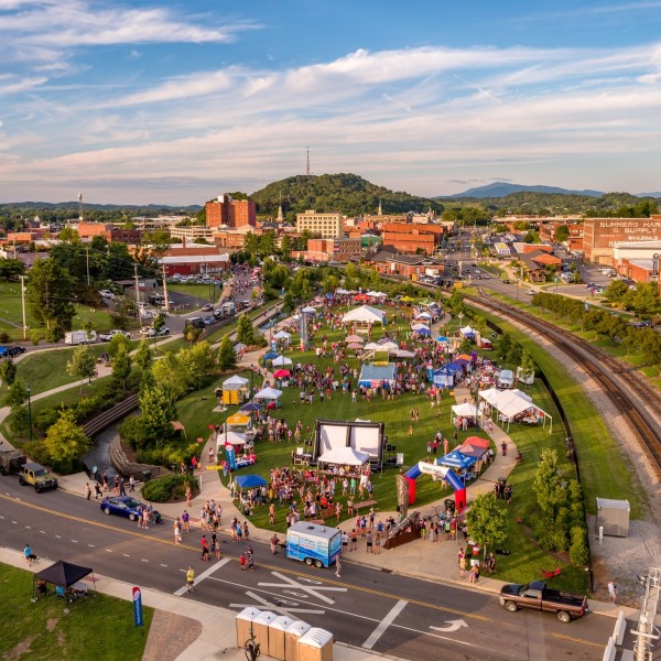 Aerial view of a community event in an open green space bordered by a road and train tracks; in the background, a downtown district is visible.