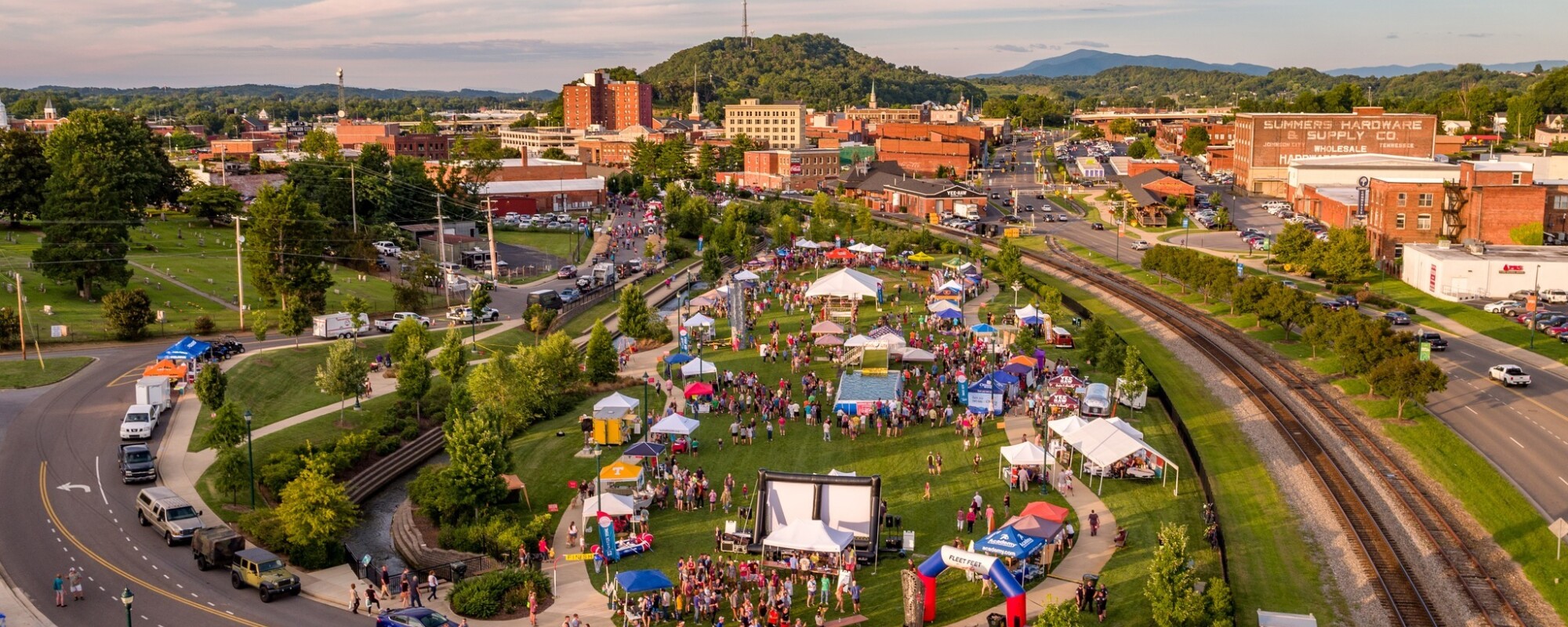 Aerial view of a community event in an open green space bordered by a road and train tracks; in the background, a downtown district is visible.