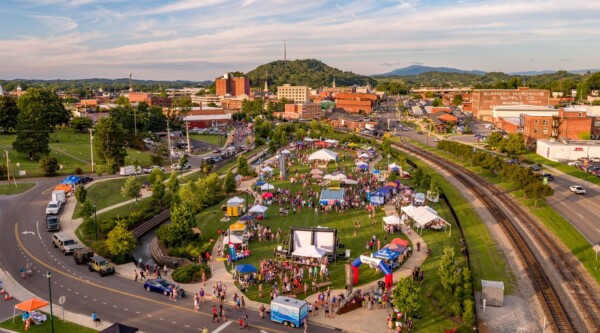 Aerial view of a community event in an open green space bordered by a road and train tracks; in the background, a downtown district is visible.