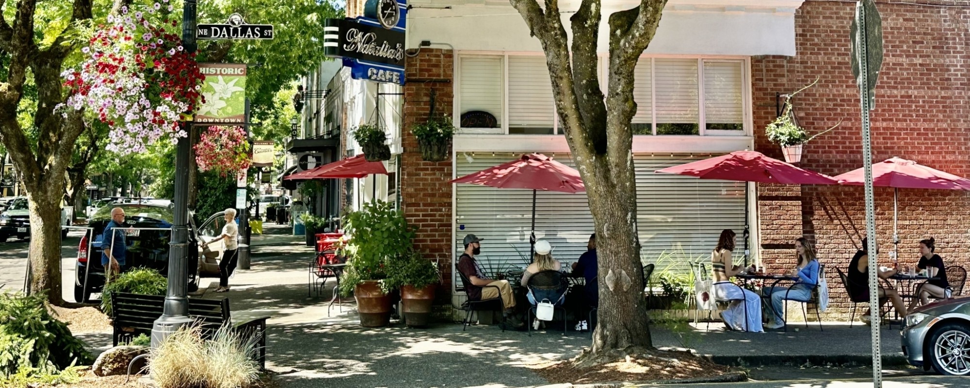On a sunny day, diners enjoy outdoor sidewalk seating under the shade of trees and meals from a café located in a historic brick building.