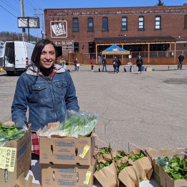 In a surface parking lot, a woman stands behind a table covered with cardboard boxes and paper bags filled with fresh produce.