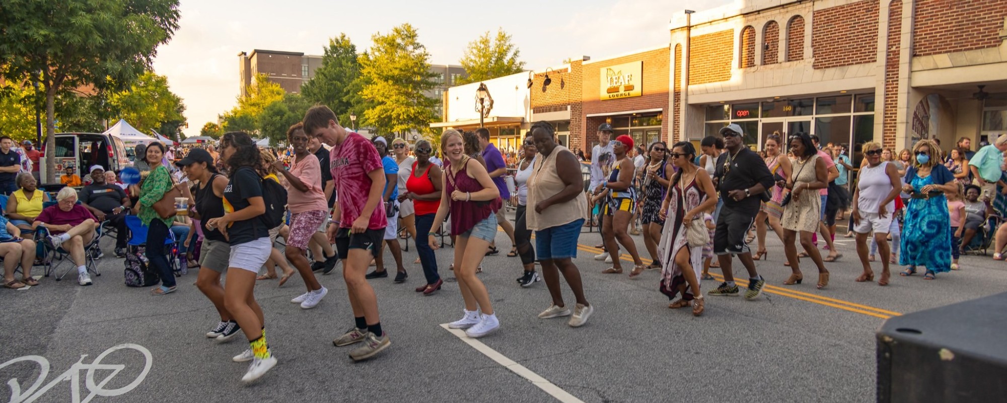 People of all ages and backgrounds line dance in the middle of a street at a summer event.
