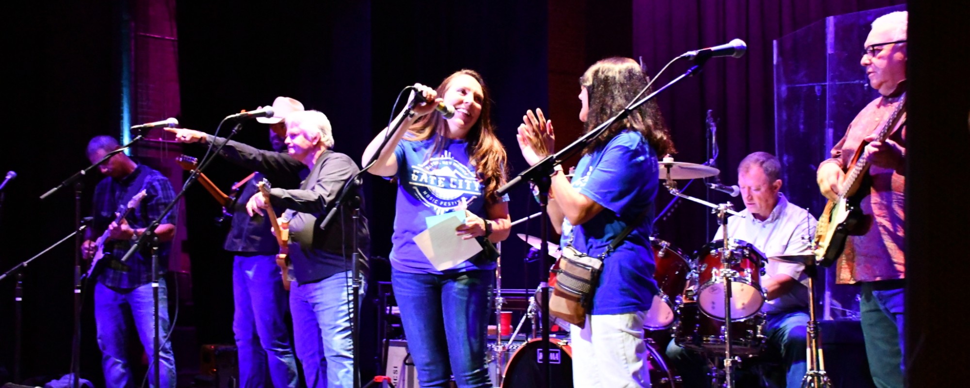 A woman stands center stage while speaking into a microphone and surrounded by a band.