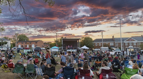 At sunset, group of people gather on lawn in front of stage.