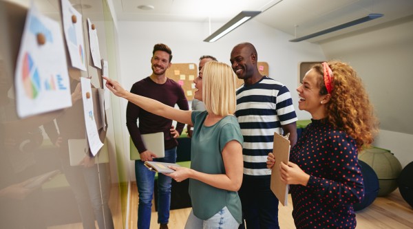 Diverse group of smiling colleagues looking at graphs and charts taped to a wall while having a meeting together in a modern office.