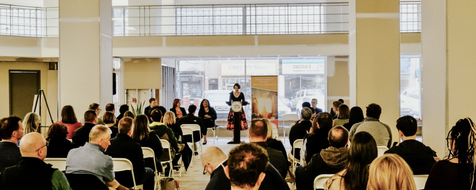 A woman stands at a podium while giving a presentation to dozens of people sitting in chairs theater style.