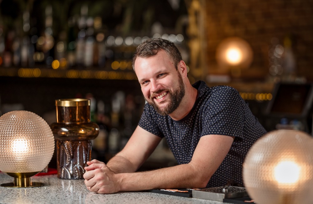 A man smiles while leaning on a marble bar top that is illuminated by globe-shaped glass lamps.