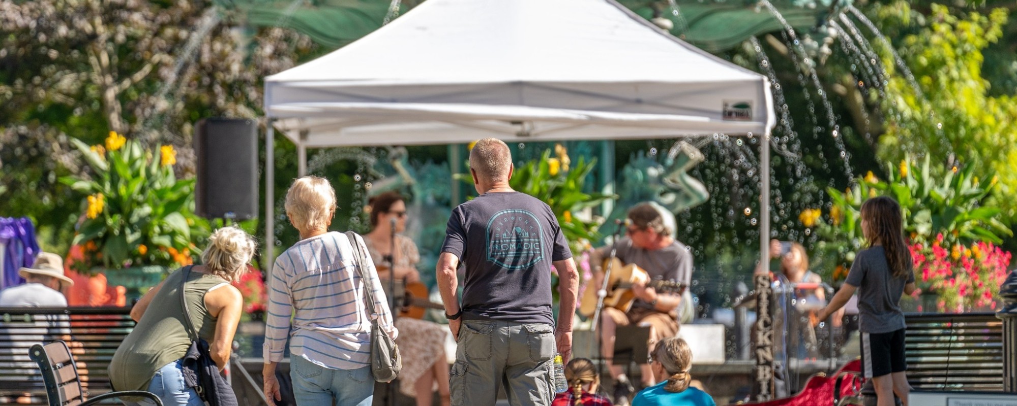 Musicians play in front of a large fountain in a park while people sit on the lawn and walk on a path.