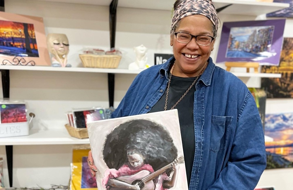 A woman smiles while holding a painting; behind her works of art are on display.