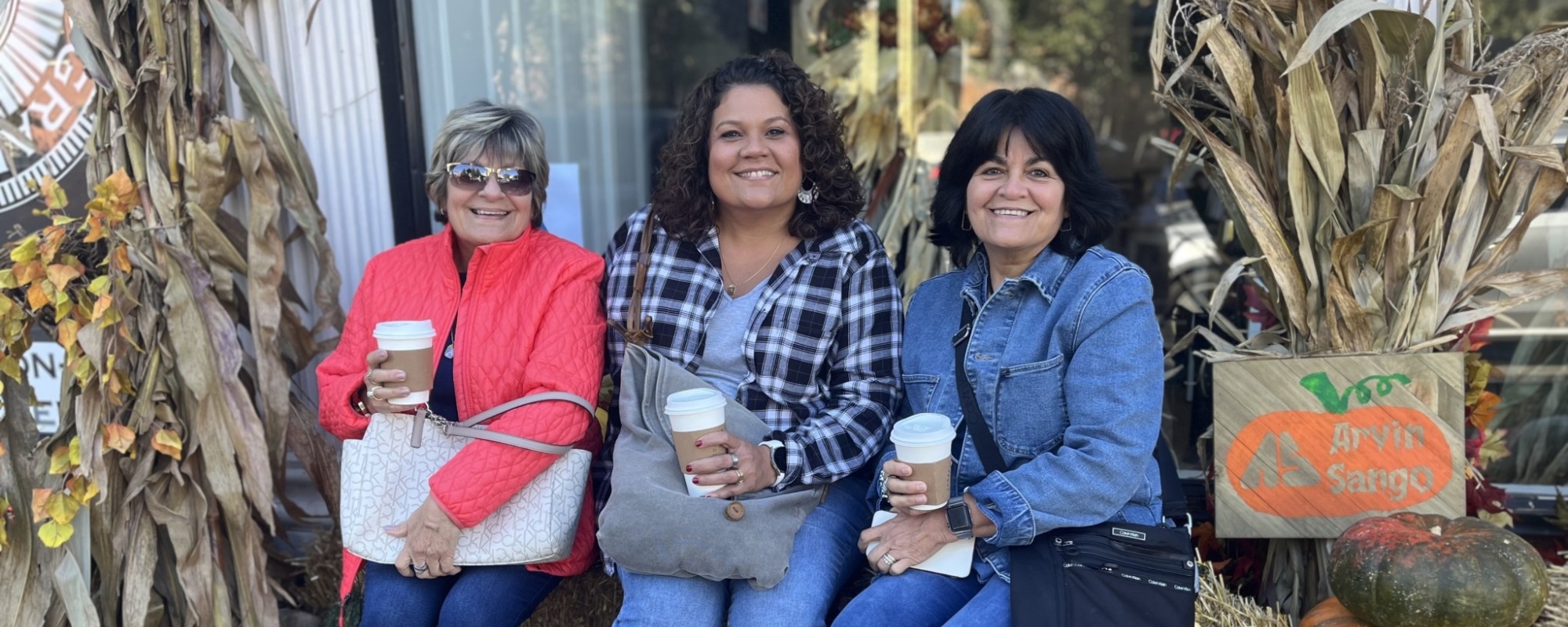 Three women hold cups of coffee while sitting on a hay bale.