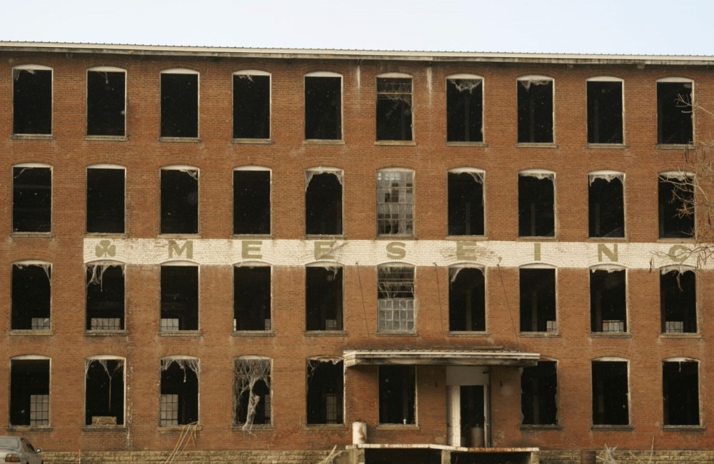 Large, historic mill building with deteriorated sign and broken windows.