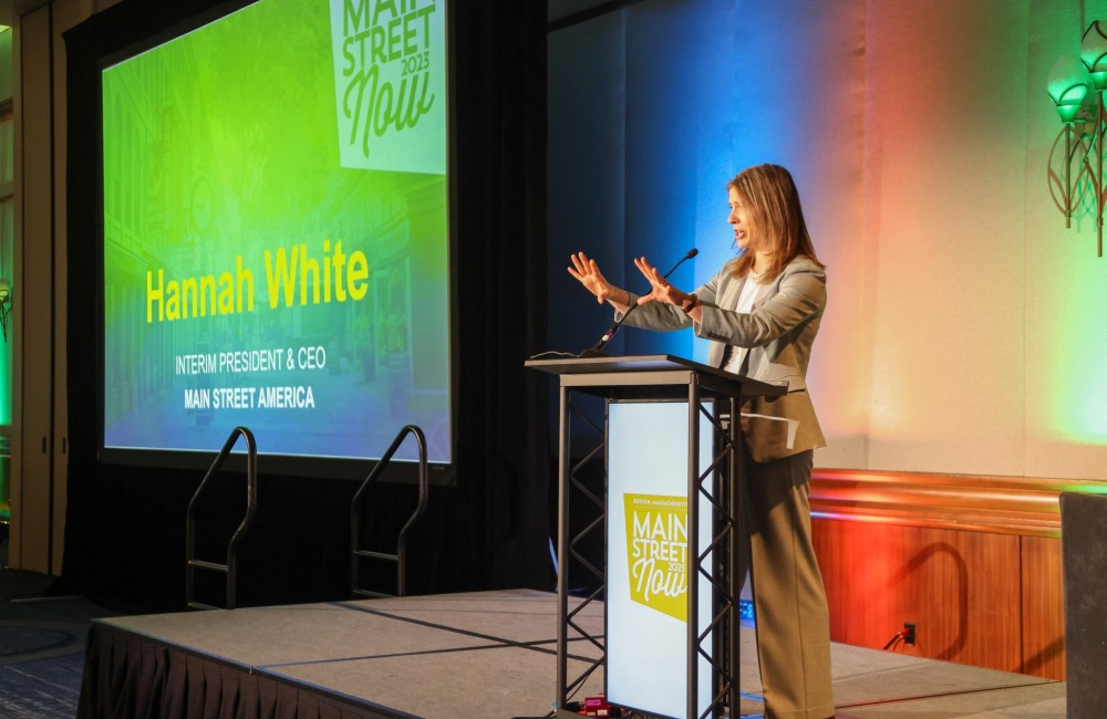 A woman gestures with her hands while speaking at a podium on stage; a large screen to her right displays slides.