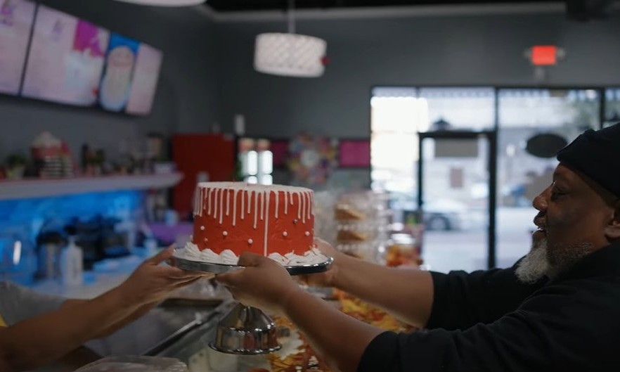 Photo of older woman with glasses handing a decorative pink cake to an older man with a beard wearing a black beanie. They're standing over a bakery counter in a small business.