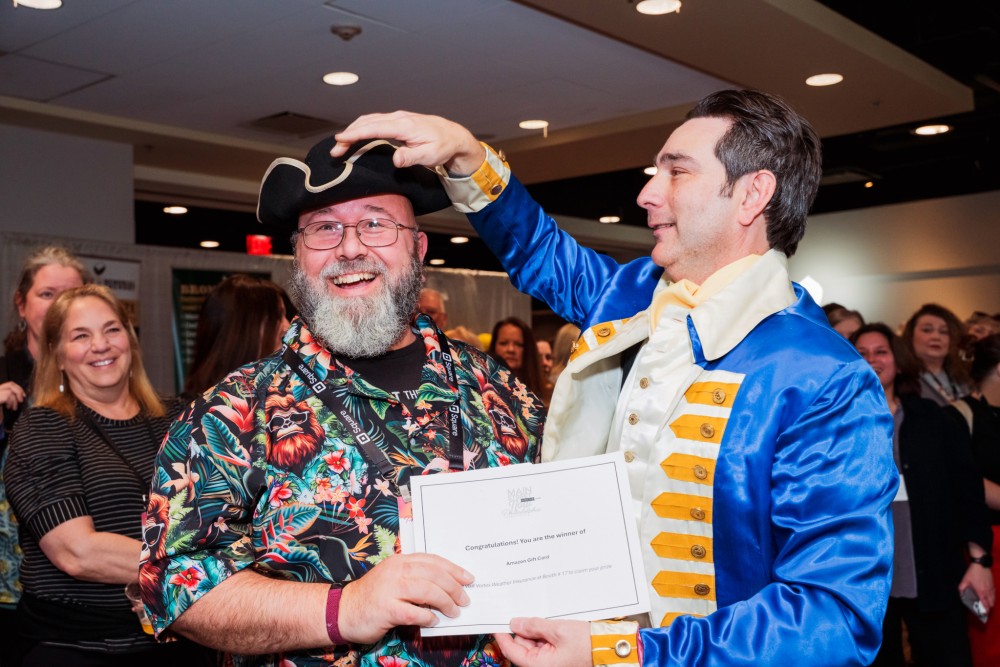 A man smiles while another man dressed in American Revolutionary War-style garb places a tripoint hat on his head and hands him a gift certificate.