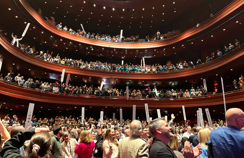 A large theater filled with people, some holding signs displaying the names of US states.