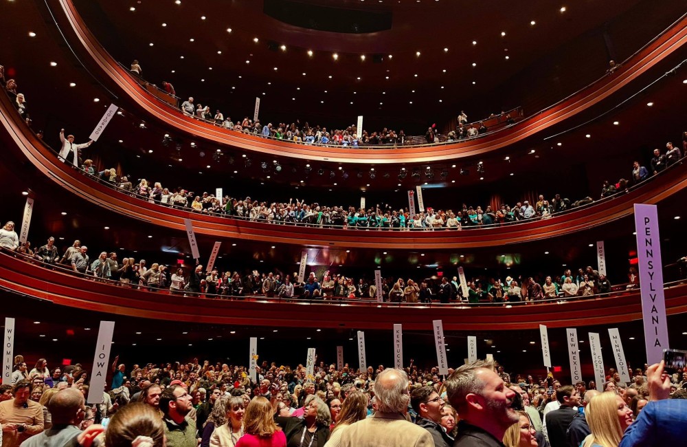 A large theater filled with people, some holding signs displaying the names of US states.