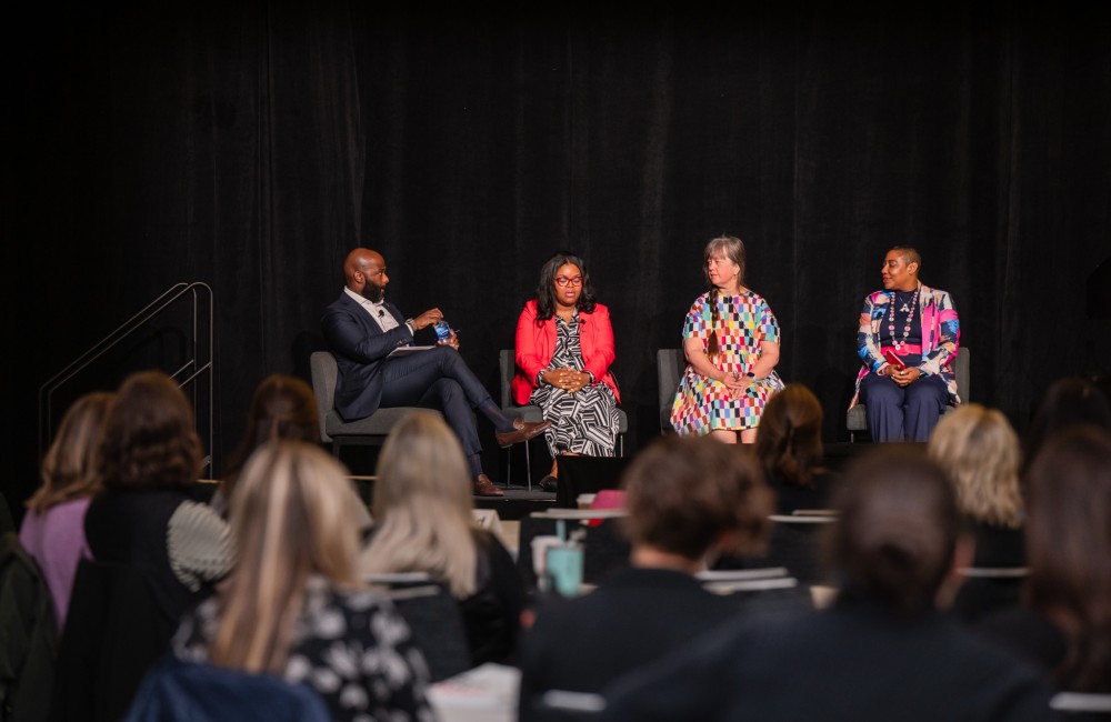 Four people sit in chairs on a stage; the three people on the right look at the person on the left who is speaking.
