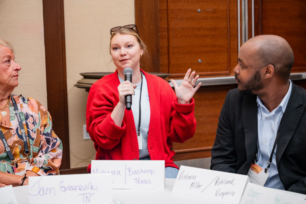 A woman, sitting at a table with other people, speaks into a microphone.