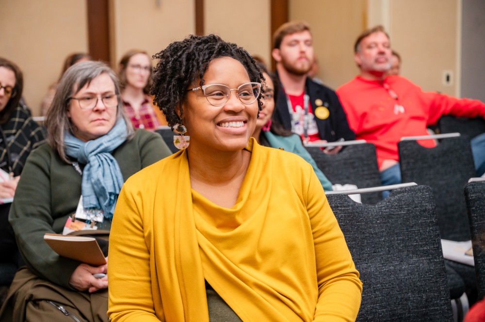 People sitting in a conference room listen intently to a presentation; in the foreground, a woman smiles.