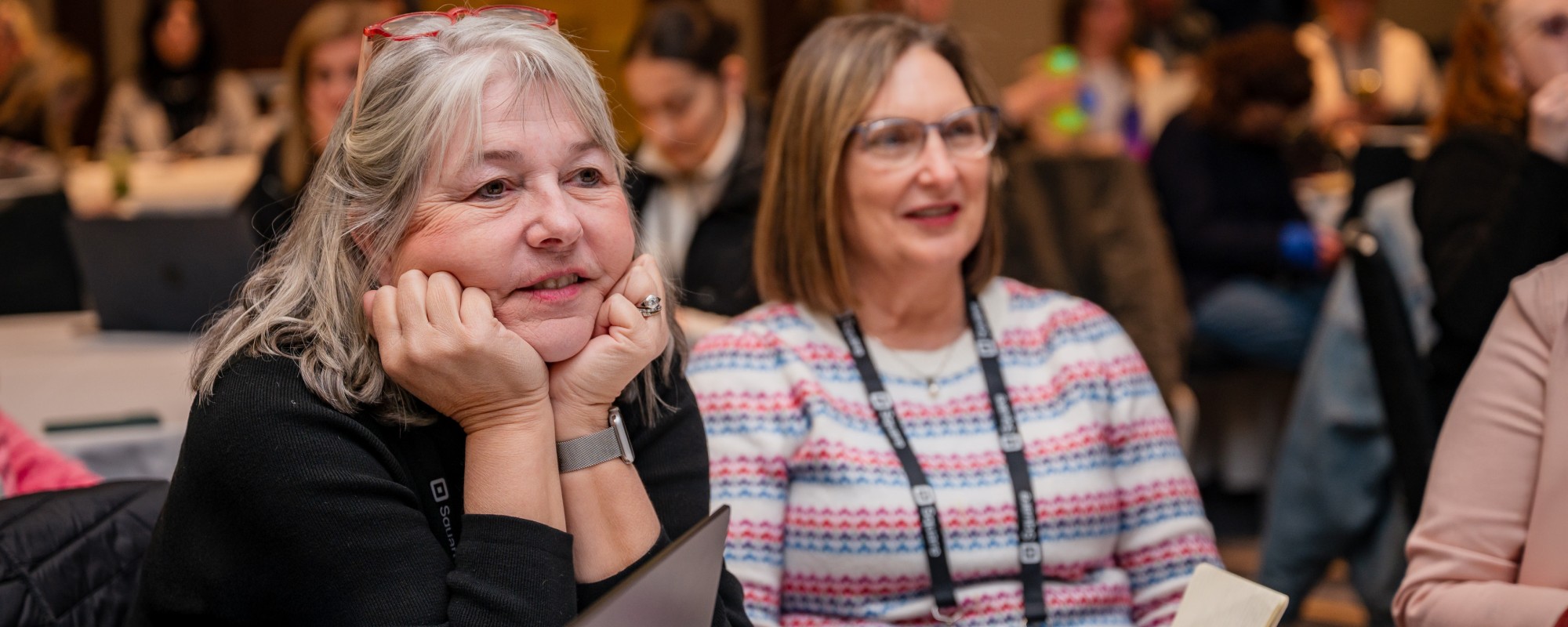 Two women are sitting at a table and listening intently to a speaker.