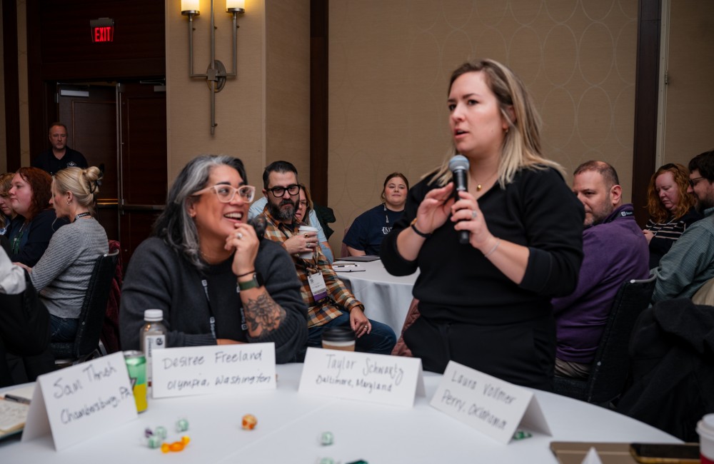 A woman speaks into a microphone while standing in a room filled with people sitting at round tables.