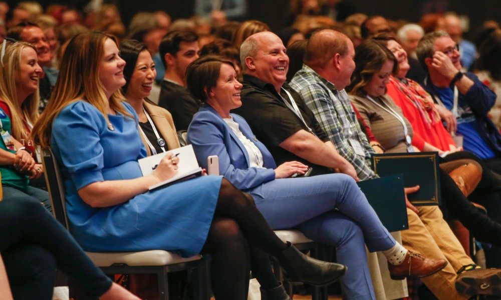 People sitting theater-style in a large room, laughing along with a speaker (not pictured).