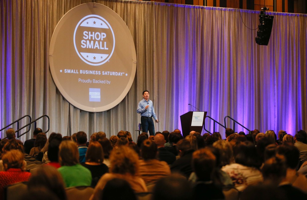 A person on stage speaks to crowd people seated in rows.