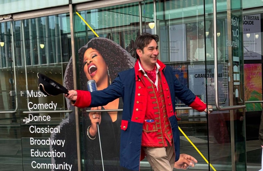 A man dressed in an American Revolutionary War coat smiles while holding a door open for guests.