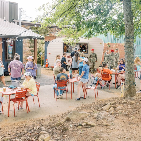 In an outdoor public seating area, people line up to place orders at a coffee shop operating from a small vintage-style travel trailer, while others enjoy their beverages while sitting at tables.
