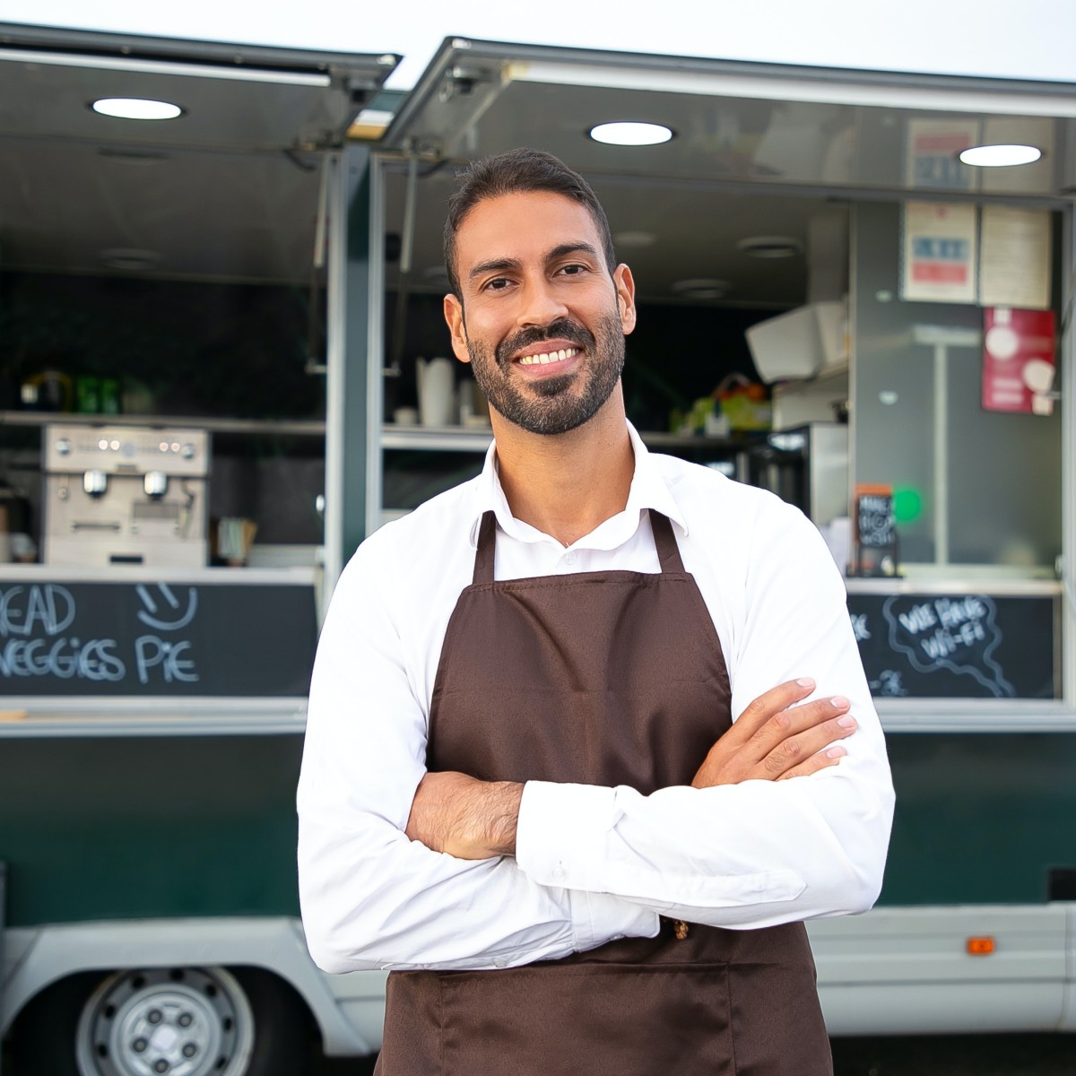 A man in a brown apron crosses his arms and smiles, he stands in front of a green food truck.