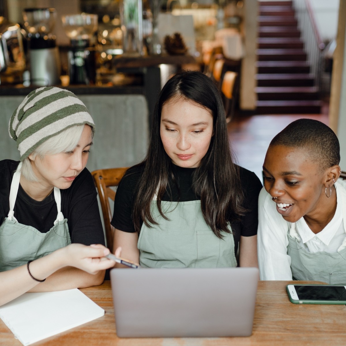 A group of three women in aprons gather around a computer in a coffee shop.