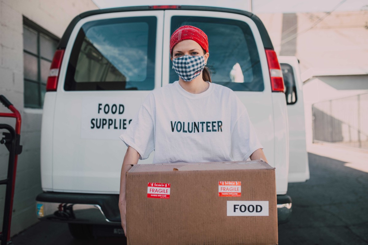 A woman volunteer with a red headband and face mask holds a cardboard box labeled "food" in front of a white van labeled "food supplies."
