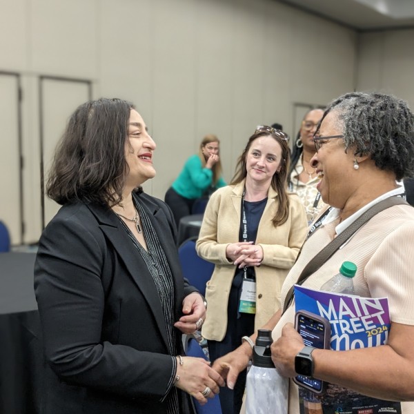 Two women shake hands and chat in a room filled with people networking.
