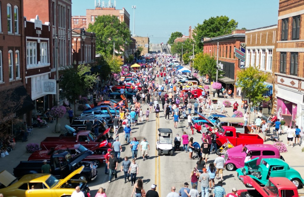 Aerial view of a wide street lined with classic cars on display and historic brick buildings; hundreds of people stroll throughout the space on a sunny day.